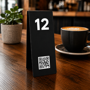 Black stand with a table number sign 12 and QR code on a wooden table with a coffee cup and plant in the background