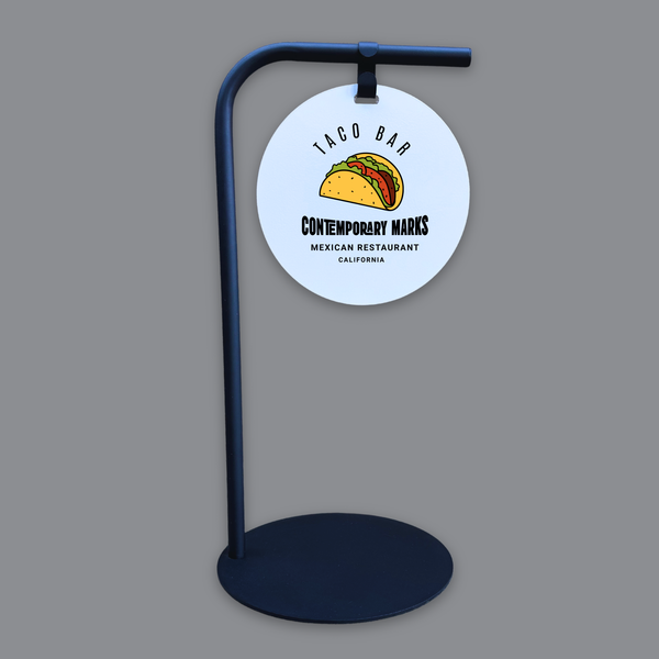 Circular sign with taco illustration and text on a stand against a gray background