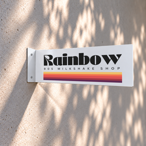 A white rectangular wall sign for 'Rainbow 90s Milkshake Shop' mounted on a textured beige wall, with dappled shadows cast across it. The sign features bold black lettering and a retro horizontal stripe pattern in orange, yellow, and purple.