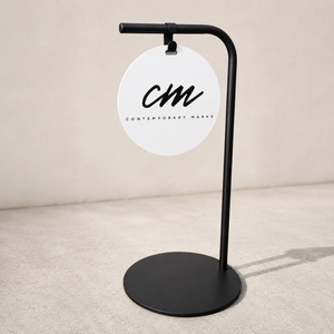 Black sidewalk sign with a white circular sign displaying 'CM Contemporary Marks' on a beige background