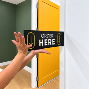 A black rectangular blade sign with yellow arrows and the words 'ORDER HERE' in white, held by a person's hands in front of a bright yellow door in an indoor room.