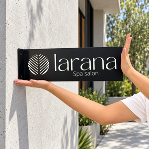 A black projecting metal custom sign for 'larana Spa salon' with a white leaf logo, being held against a light gray concrete wall by a person's hands and arms.