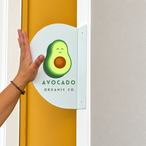 A white half round sign featuring a smiling avocado graphic and the text 'AVOCADO ORGANIC CO.' against a mustard yellow door, with a light gray wall on the right.