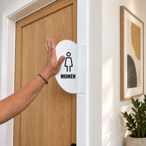 A white oval restroom sign with a black female icon and the word 'WOMEN' mounted on a light wood door.