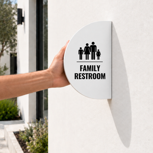 A white, semi-circular outdoor blade sign against a light beige stucco wall. The sign features a black icon of a family of five above the text 'FAMILY RESTROOM'.