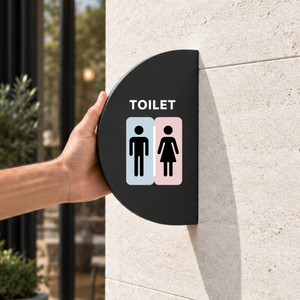 A black half round toilet blade sign mounted on a beige stone wall. The sign features the word 'TOILET' above male and female restroom icons on light blue and light pink backgrounds.