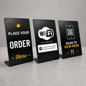Three large black metal table countertop signs displayed on a gray surface against a gray background. The left sign says 'PLACE YOUR ORDER Here' with a yellow bell icon, the middle sign displays 'Wi Fi' card graphic, and the right sign says 'SCAN TO VIEW MENU' with a QR code and fork and knife icons.