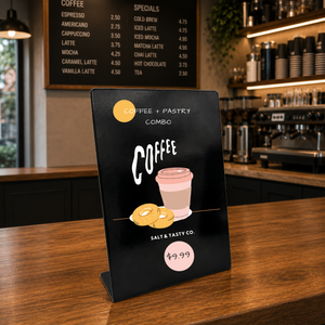 A black tabletop coffee combo menu sign from Salt & Tasty Co. stands on a wooden counter in a cafe setting, with a coffee menu board and bar shelves.
