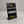 A black solid metal tabletop sign with a metal stand, featuring customizable white and yellow text that reads 'PLACE YOUR ORDER' and 'Here', set against a neutral gray background.