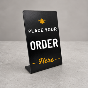 A black solid metal tabletop sign with a metal stand, featuring customizable white and yellow text that reads 'PLACE YOUR ORDER' and 'Here', set against a neutral gray background.