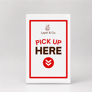 A white rectangular table sign or with a red border and the text 'Layer & Co.' and 'PICK UP HERE' in red and brown, plus a red circular chevron icon.