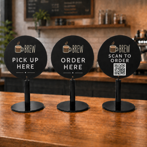 Three round black coffee shop round table signs displayed on a wooden counter in a cafe setting. The signs read 'BREW PICK UP HERE', 'BREW ORDER HERE', and 'BREW SCAN TO ORDER' with a QR code on the right.