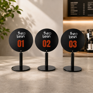 Three round black tabletop signs on a countertop in a cafe setting. Each sign displays custom logo in white script with a large orange number underneath: 01, 02, and 03.