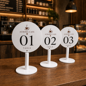 Three round white table numbers signs in 01, 02, and 03 sequence, printed with 'AURORA CAFE TABLE' and a coffee cup icon, displayed on a wooden table in a cafe setting.