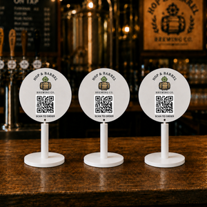 Three white circular bar table signs with QR codes and the Hop & Barrel Brewing Co. logo, displayed on a dark wooden bar counter in a bar setting.