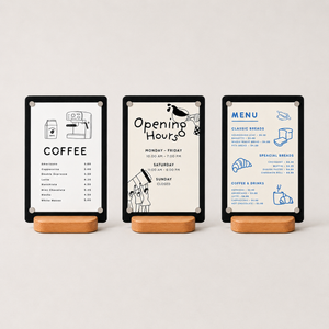 Three black table menu sign displayed on wooden stands.  The signs feature clean minimalist typography and line drawings: one coffee menu, one opening hours notice, and one bread and coffee menu.