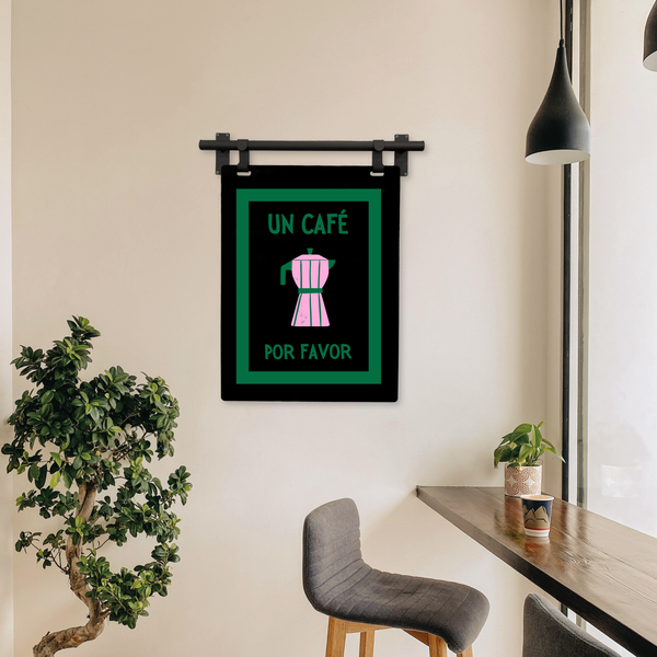 Framed artwork with 'Un Cafe Por Favor' text and coffee pot illustration on a wall in a room with plants and a chair.